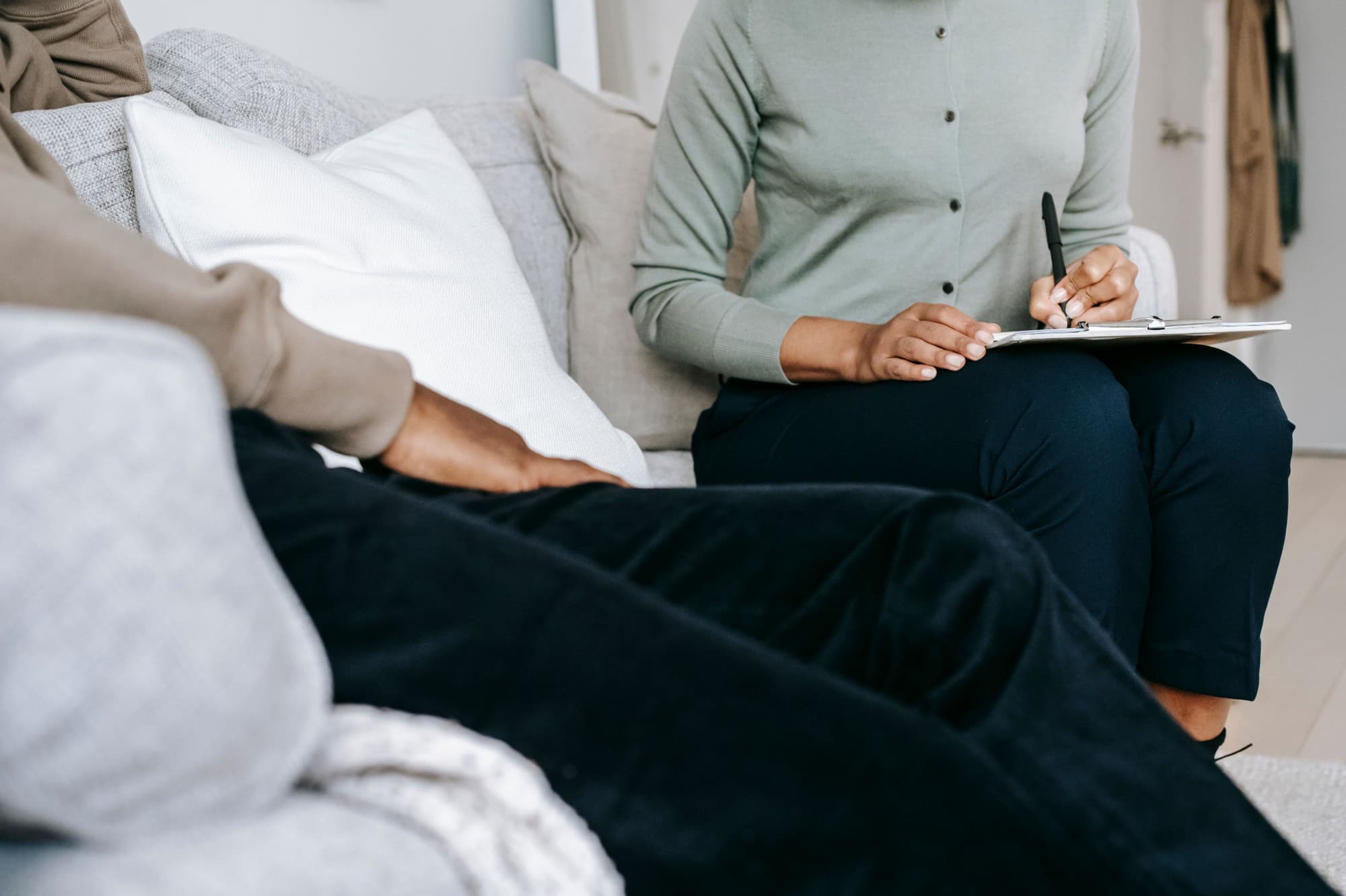 A clinician taking notes on a clipboard while a client talks from the sofa