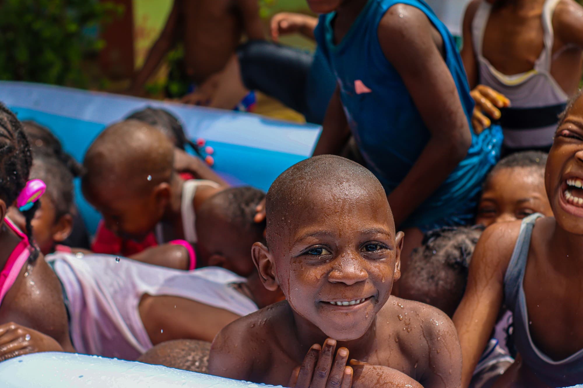 A smiling child resting his chin on the edge of a paddling pool, with friends playing behind him
